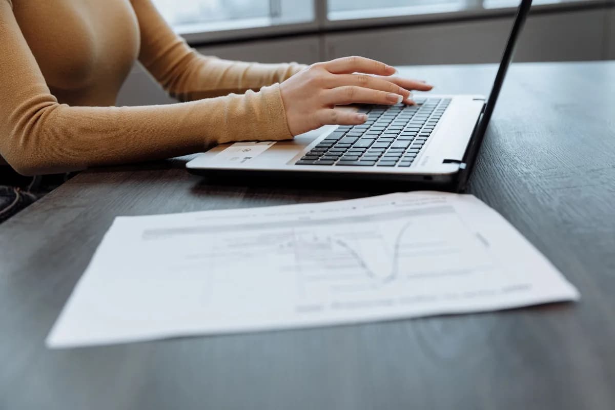 Mãos digitam em um notebook sobre uma mesa escura, ao lado de um documento impresso com gráficos