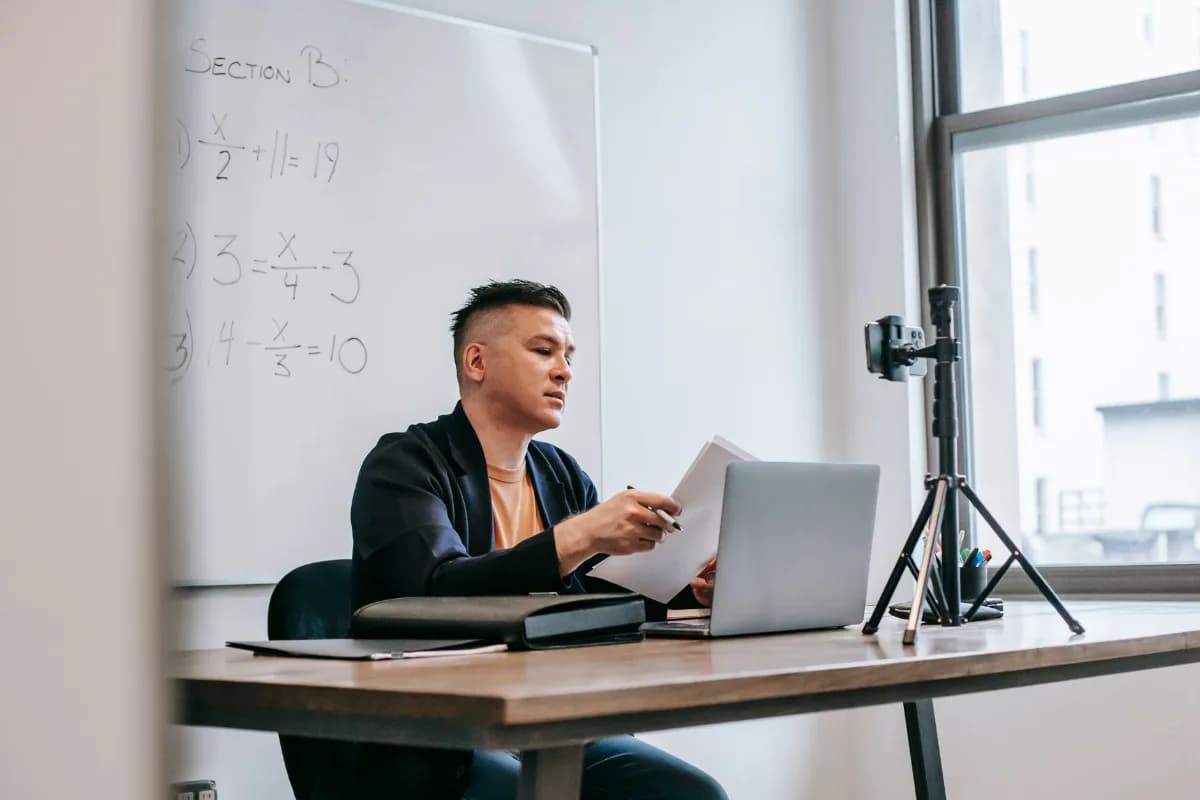 Professor em sala de aula conduz uma aula online com notebook e câmera montados à sua frente