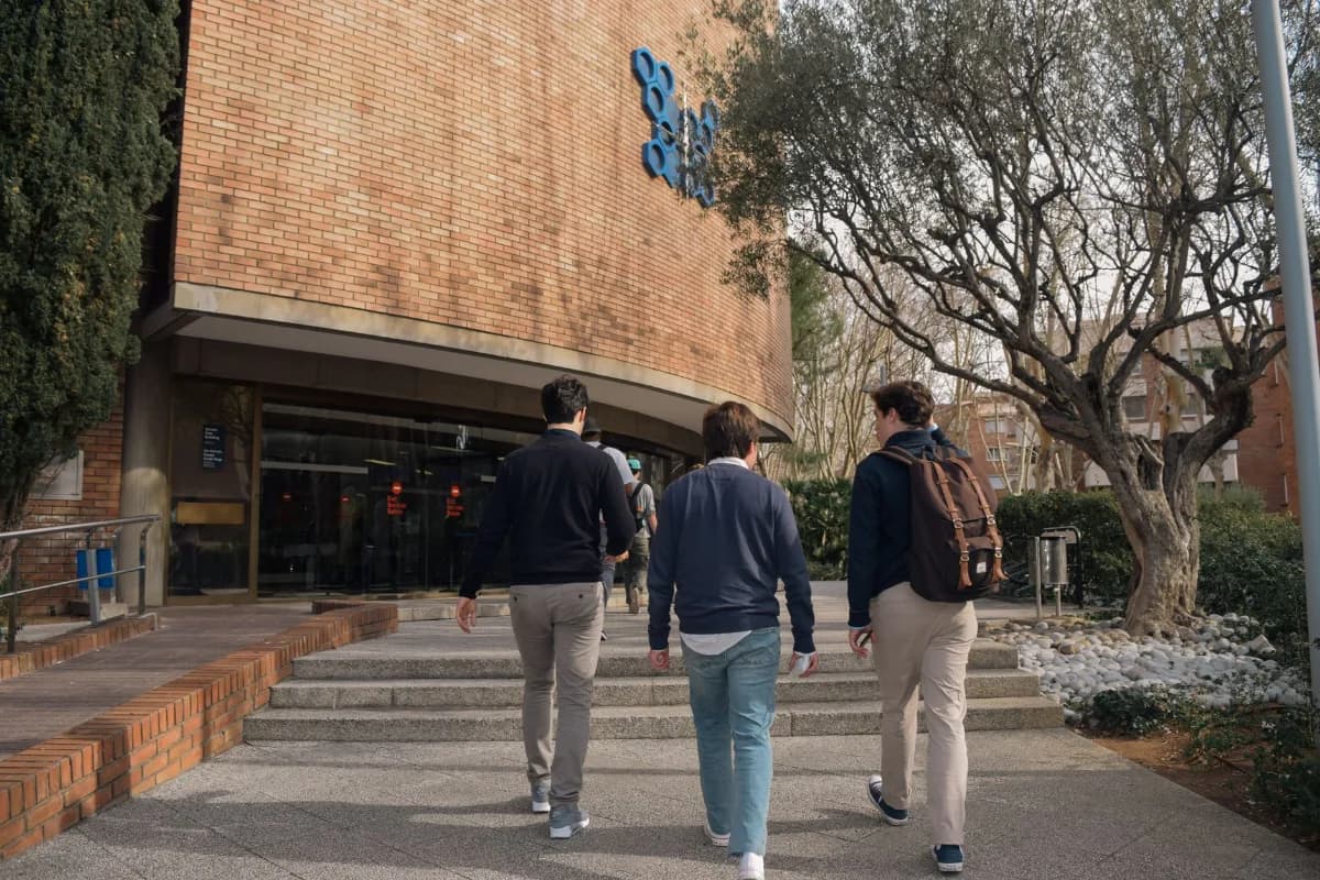 Três estudantes de costas caminham em direção à entrada de um prédio universitário de tijolos aparentes